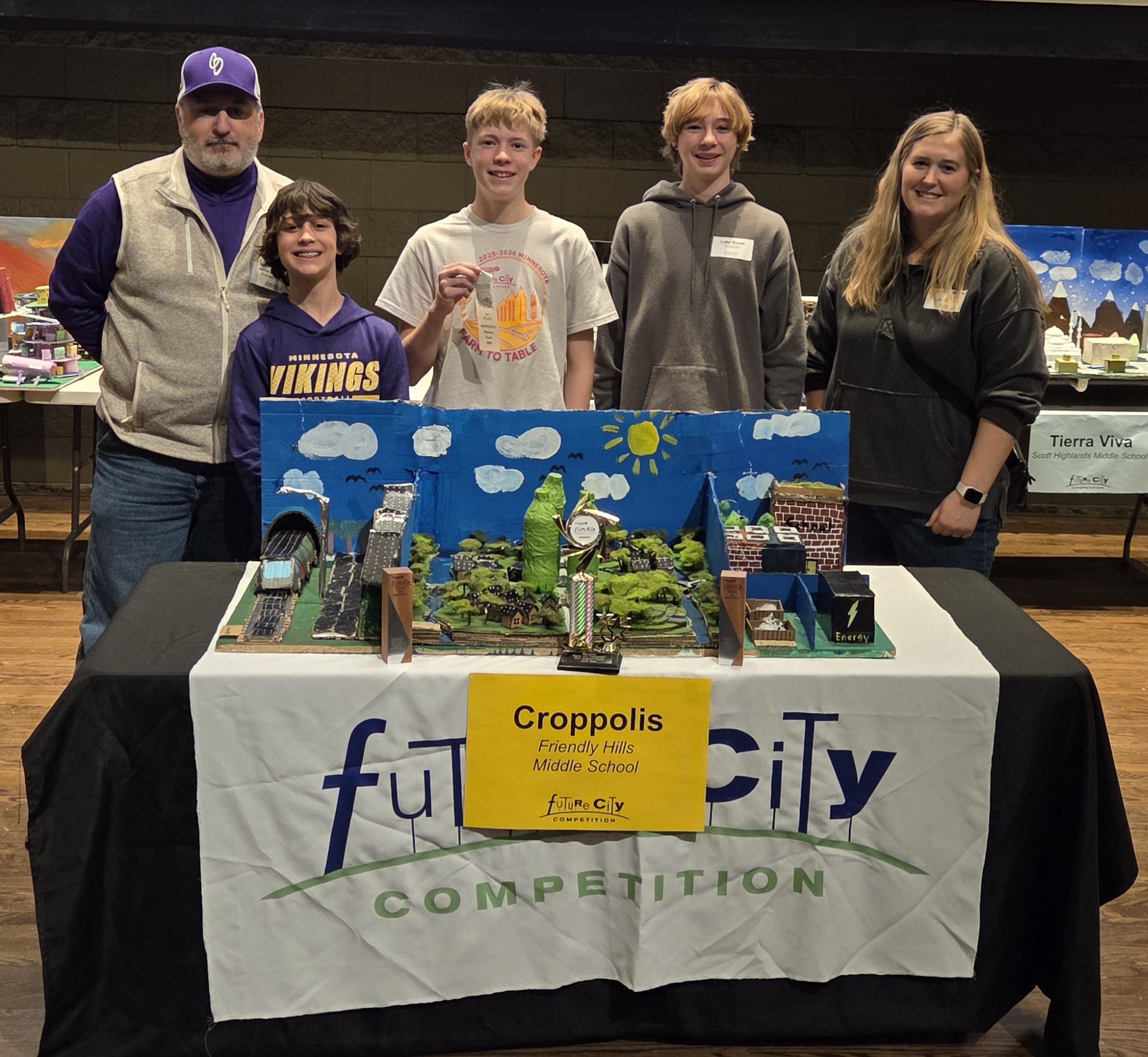 Five people stand behind a model display table for a "Future City" competition. They are smiling and posing.